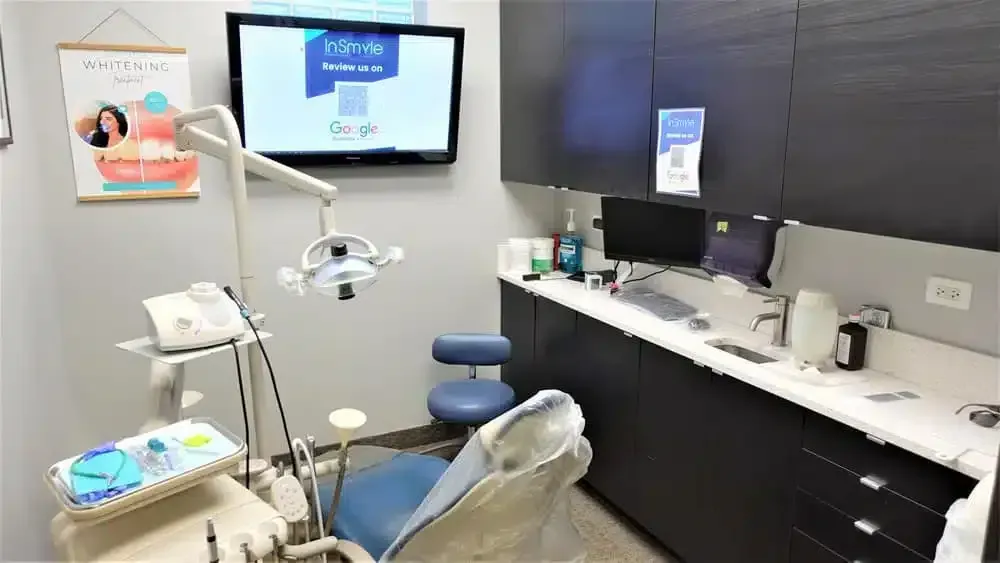 Dental office treatment room with examination chair, overhead light, monitor, and sink counter.