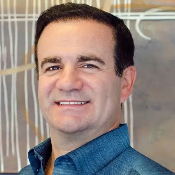Smiling man with short dark hair wearing a blue collared shirt, indoors.